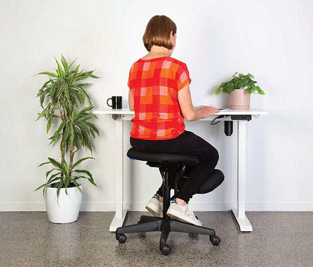 Lady in red shirt seated on Buro Knee ergonomic chair at a height adjustable desk