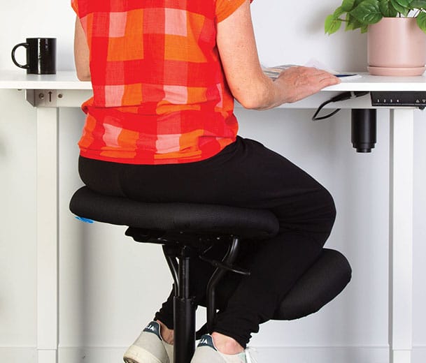 Lady in red shirt seated on Buro Knee ergonomic chair at a height adjustable desk